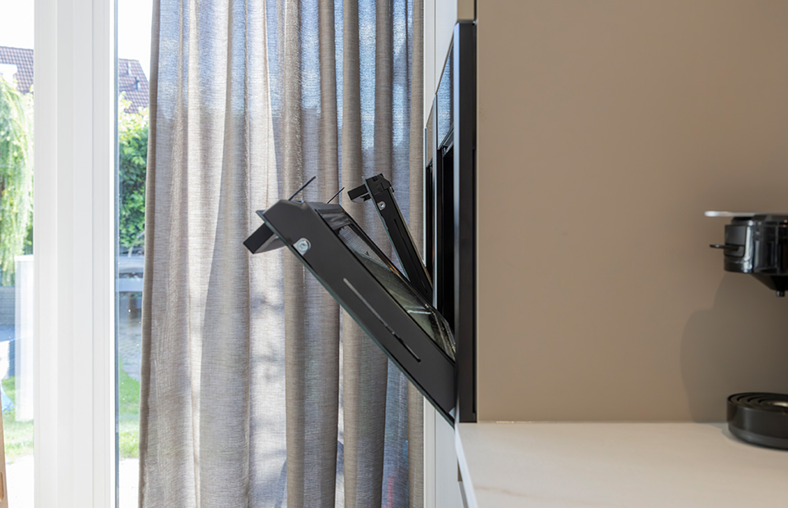 Moderne keuken met beige kasten, wit keukenblad, keukeneiland met zwarte barkrukken, hanglampen, en zicht op een tuin