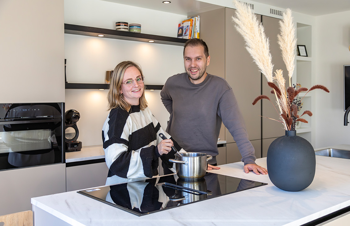 Moderne keuken met beige kasten, wit keukenblad, keukeneiland met zwarte barkrukken, hanglampen, en zicht op een tuin